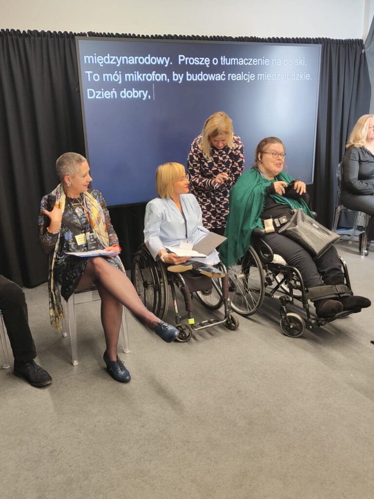 Prowadzące sesje ogólne Anna Rutz i Małgorzata Peretiatkowicz oraz witająca uczestniczki przewodnicząca Komisji kobiet w European Disability Forum. Towarzyszy in tłumaczka. Za kobietami duży ekran na nim widoczna transkrypcja.